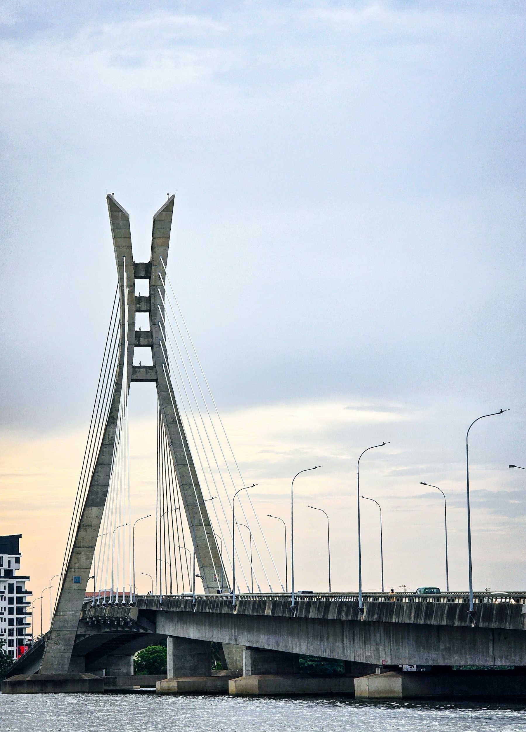 A long cable-stayed bridge spanning a wide lagoon, with Lagos skyline behind, at golden hour.